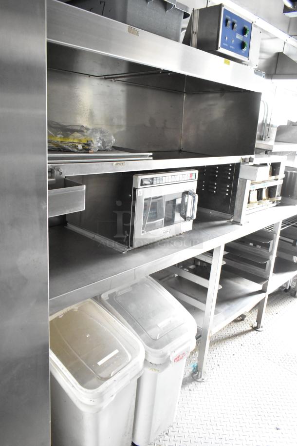 Fully outfitted food trailer interior with stainless steel shelving, industrial microwave, storage bins, and electrical control panel.