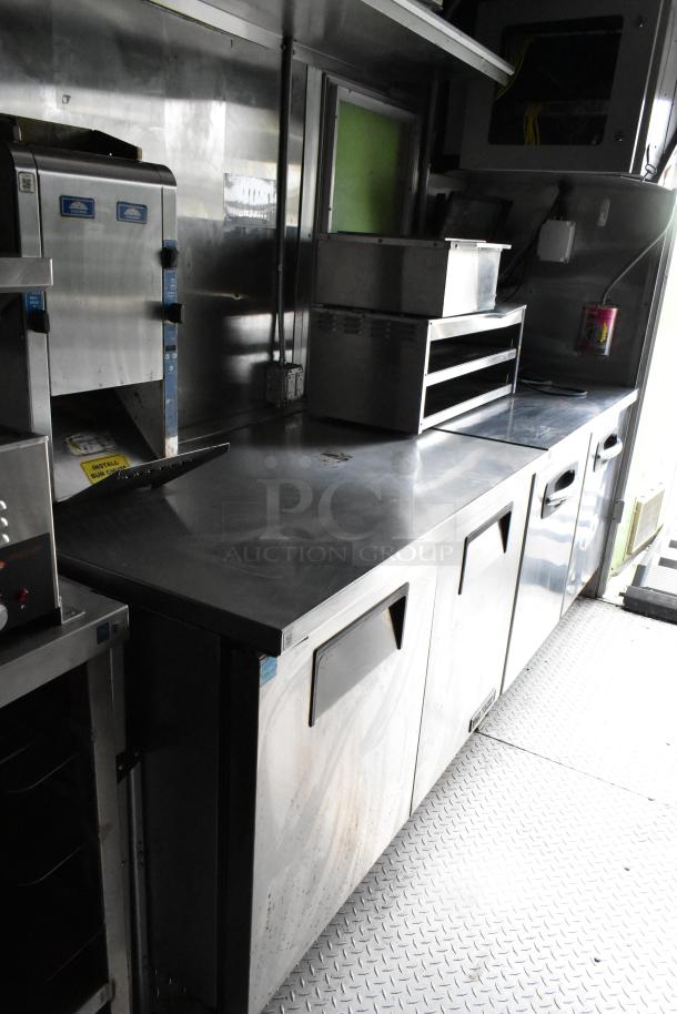 Stainless steel interior of a fully equipped food trailer with commercial appliances, work surfaces, and under-counter storage.