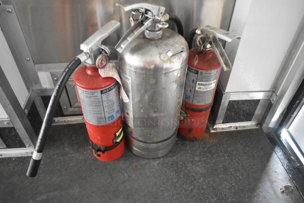 Three fire extinguishers inside an SDG trailer, showing light wear and visible instruction labels.