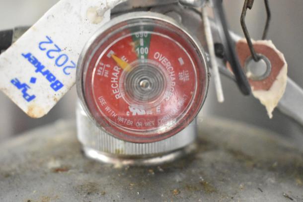 Close-up of a pressure gauge with "Recharge" and "Overcharge" zones, attached tag reads "2022." Use with water or wet chemicals.