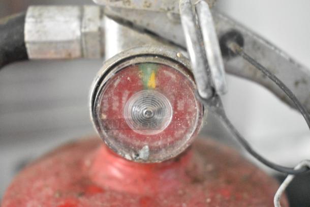Close-up of a pressure gauge on a red fire extinguisher, showing moderate wear and visible dirt on the surface.