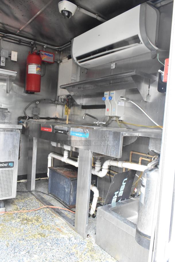 Interior of a fully outfitted food trailer with stainless steel fixtures, sink, air conditioning unit, fire extinguisher, and appliance connections.