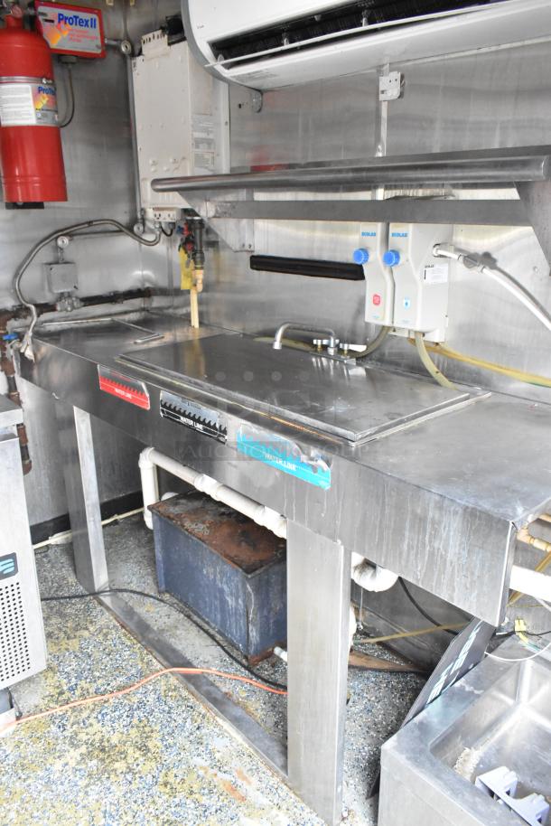 Stainless steel countertop with attached sink, labeled water lines, and ProTex II fire suppression system in food trailer.