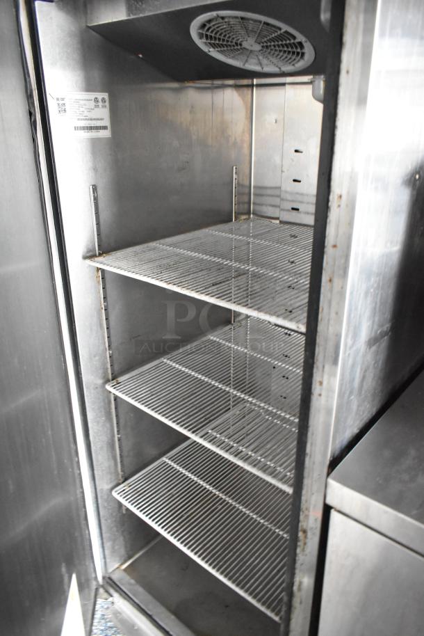 Commercial fridge interior with metal shelves, cooling fan at top. Label with barcode and safety symbols on left wall.