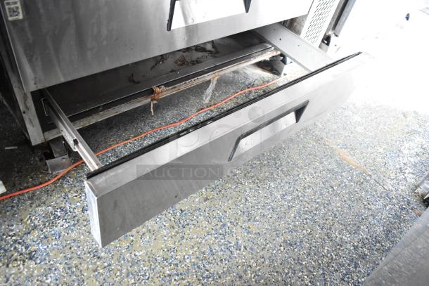 Stainless steel drawer from a food trailer appliance, slightly open, showing wear. Visible orange cable on speckled floor.
