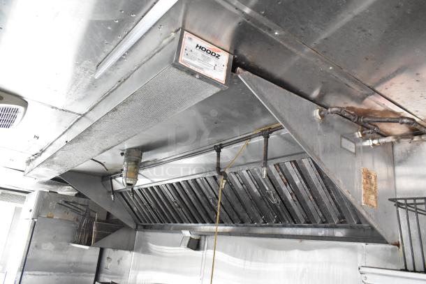 Stainless steel kitchen exhaust hood in a food trailer, featuring a Hoodz cleaning label, ductwork, and overhead lighting.