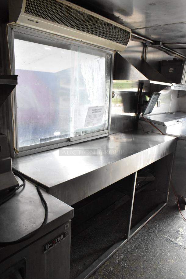 Interior view of a fully equipped food trailer with stainless steel countertops, ventilation hood, and appliances. Visible label on the equipment.