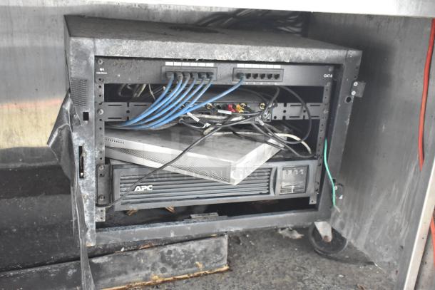 Server rack with multiple blue CAT6 cables and APC unit in metal enclosure, visible wear and tear.