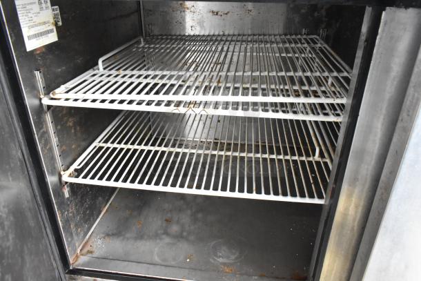 Interior view of food trailer fridge with three empty wire racks and some visible wear and rust. Labels on door indicate specifications.