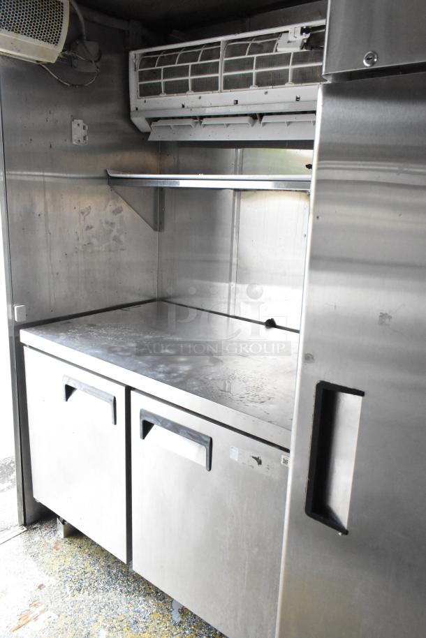 Food trailer interior: stainless steel counter, shelving, and storage, under AC unit; clean and ready for use.