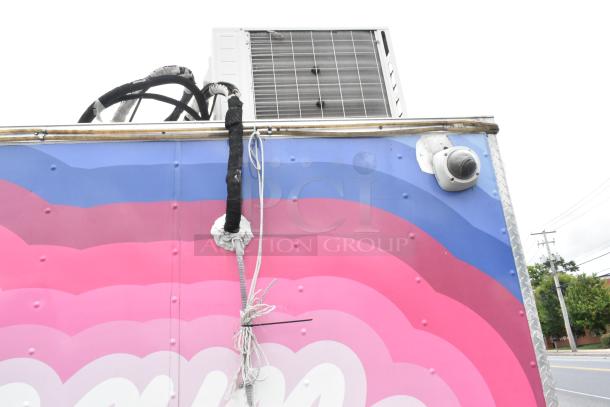 Colorful food trailer with air conditioning unit and security camera. Features vibrant pink and blue design.