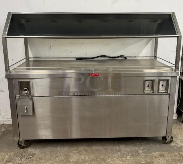 Stainless steel heated display case with glass top, control panel, and caution tag. Used condition, wheeled base.