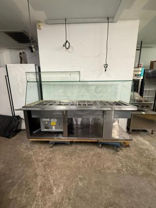 Stainless steel 12-pan steam table with glass guard, brand "Steam TAble," in used condition, on wheels for mobility.