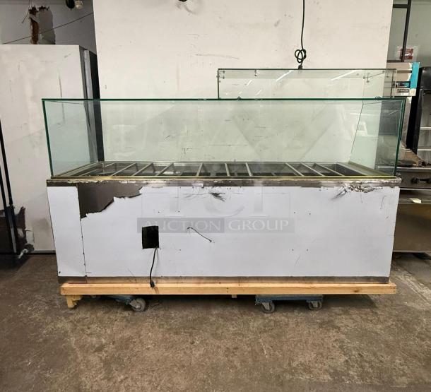 Alt text: Stainless steel 12-pan steam table with glass sneeze guard. Visible wear and scratches on the exterior. Placed on a wheeled platform.