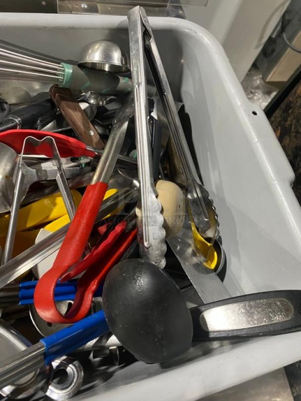 Assorted commercial kitchenware and utensils in a white bin, including tongs, ladles, and scoops. Mixed materials, used condition.