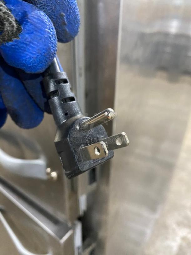 Traulsen AHT132DUTHHS refrigerator power plug held by a gloved hand, silver door with handle visible.
