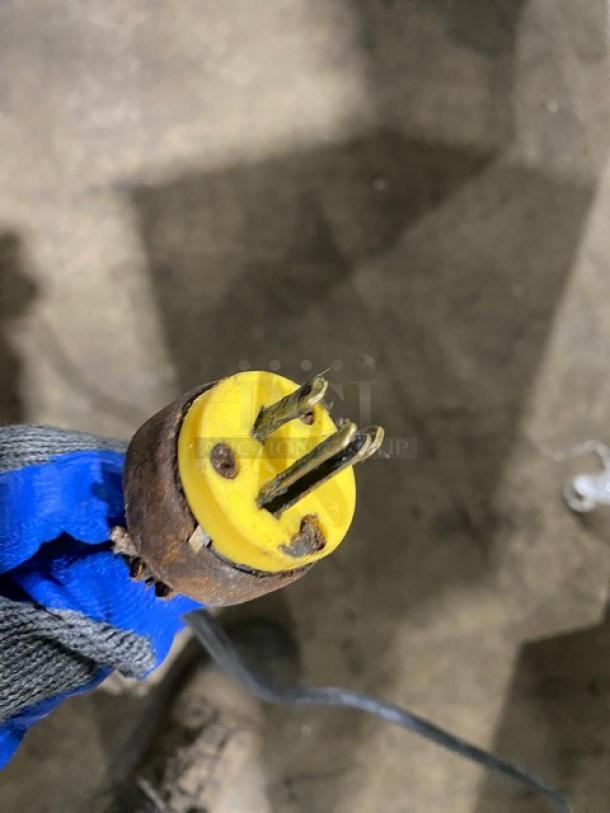 Rusted electrical plug held with gloved hand. Plug features three prongs and a yellow base. Worn condition.