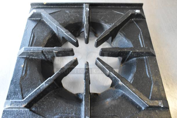 Brand new 12"x12" cast iron spider grate with open top and built-in bowl, some scratches and dents visible.