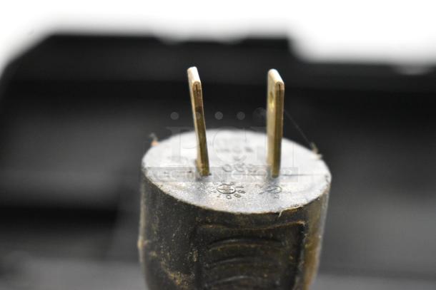 Close-up of an electrical plug showing metal prongs and circular markings. Background is a blurred tool box.