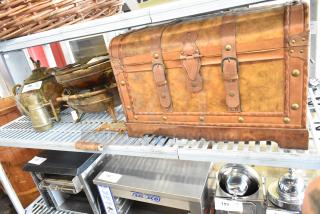 Wooden chest with leather straps and brass accents, alongside vintage brass bowls and decorative items on shelves.