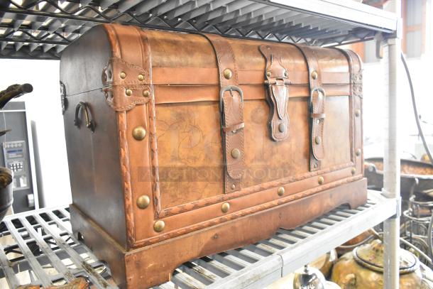Vintage leather-bound chest with brass rivets and buckles, featuring a distressed finish, stored on a metal shelf.