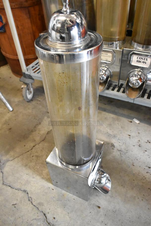 Stainless steel metal dry dispenser with visible wear, cylindrical design, clear chamber, and silver base with nozzle.