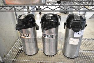 Three stainless steel airpots with black lids, labeled "Regular," "Hot Water," and "Amaretto," on metal shelf; slight wear visible.