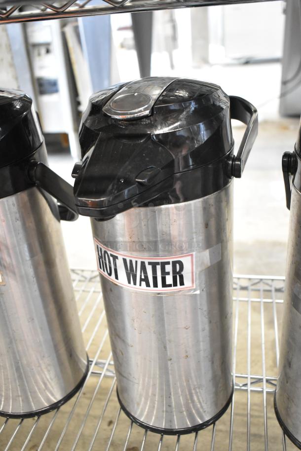 Three metal air pots labeled "Hot Water"; stainless steel with black lids, showing signs of wear, on a metal shelf.