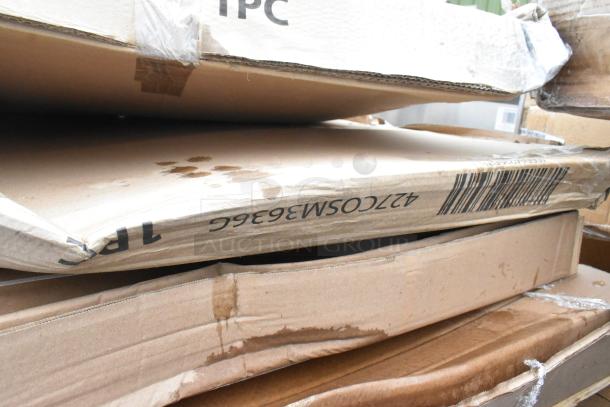 Stack of sealed boxes, marked "427COSM3636G," containing Lancaster Table & Seating Harbor Gray 36" outdoor tables. Boxes show signs of damage.