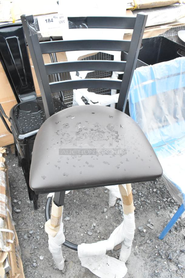 Black metal bar-height chair with ladder back, visibly scratched. Part of a lot with chairs, some wrapped, on gravel surface.