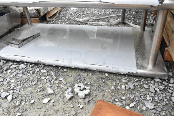 Stainless steel 2-tier shelf with visible dents and scratches, resting on gravel. Brand new condition with cosmetic damage.