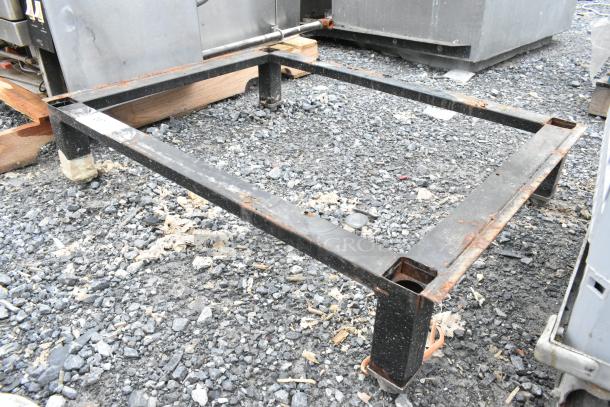 Black metal stand with rust spots, sturdy frame, and four legs. Used condition on gravel surface.