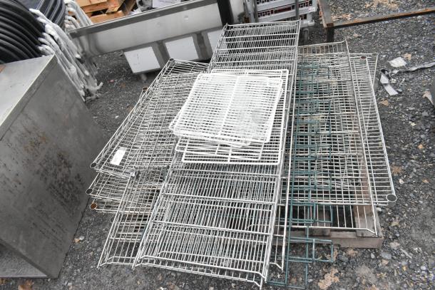 Lot of various wire shelves in mixed condition, stacked on gravel. Includes metal and coated shelves of different sizes.