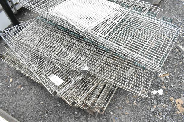 Lot of various metal wire shelves on gravel. Mixed sizes, some with green edges. Includes a small label with pricing detail.