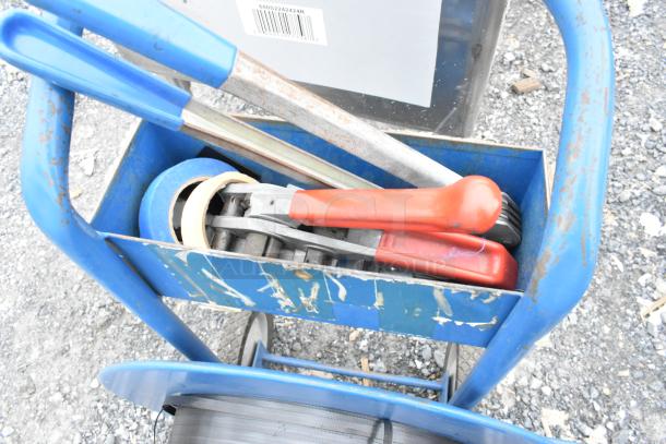 Blue metal pallet banding cart with tools and tape, showing signs of wear. Includes red-handled cutter and blue tape roll.