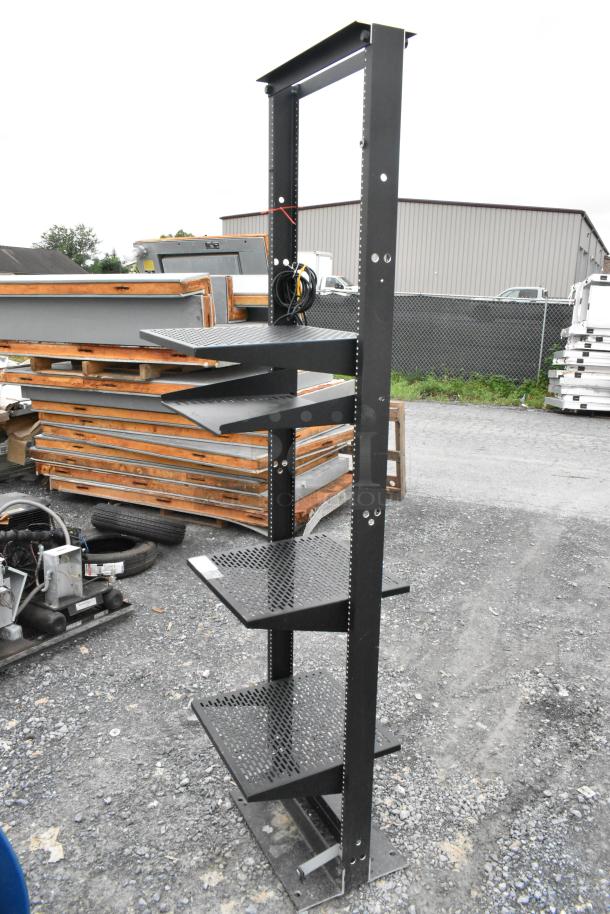 Black metal shelving unit with four perforated shelves, sturdy frame, outdoor setting, slight wear, ideal for storage.