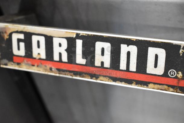 Garland brand label on a commercial gas-powered griddle with visible wear, featuring a red stripe and bold white lettering.