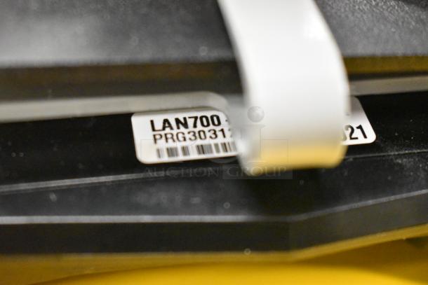 Close-up of a device with a label reading "LAN700 PRG30312" and a barcode, part of a lot including IBM and Toshiba monitors and a credit card reader.