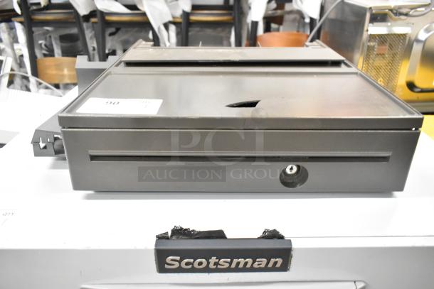 Gray metal cash drawer, featuring a lock and key slot. Minor surface wear, indicating use. Label with number 90 attached.