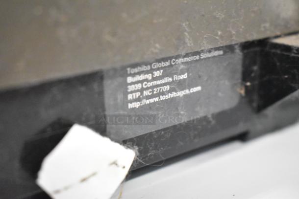 Gray metal cash drawer with visible Toshiba Global Commerce label. Shows address and website. Used condition, slight wear.