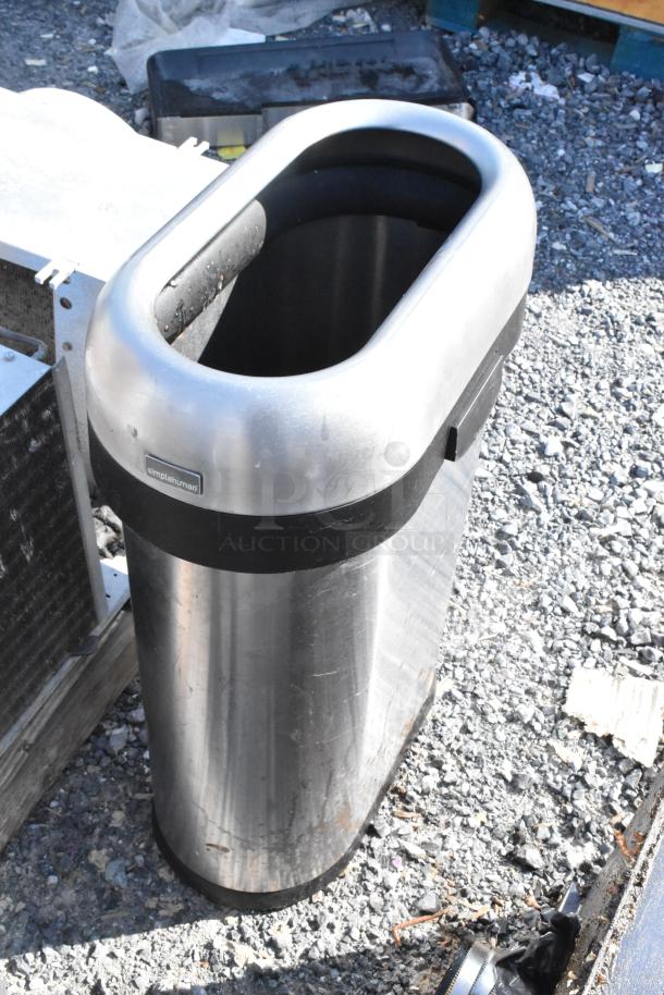 Simplehuman metal trash can, oval opening, outdoor setting, scuffed surface, black band detail; durable and sleek design.