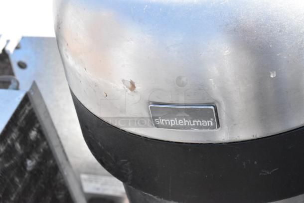Simplehuman metal trash can with visible wear and scratches, featuring a logo plate. Condition indicates prior use.