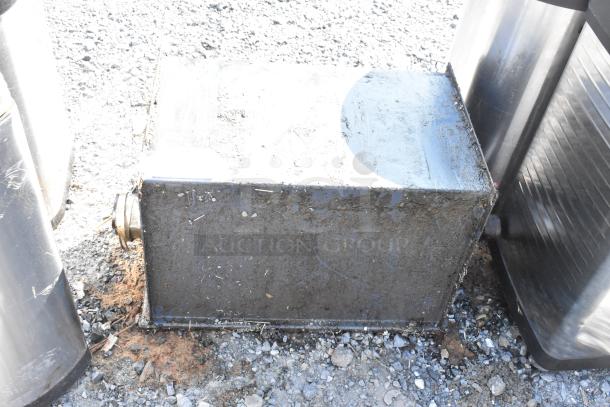 Metal commercial grease trap on gravel, showing signs of wear. Features include metal construction with a side outlet.