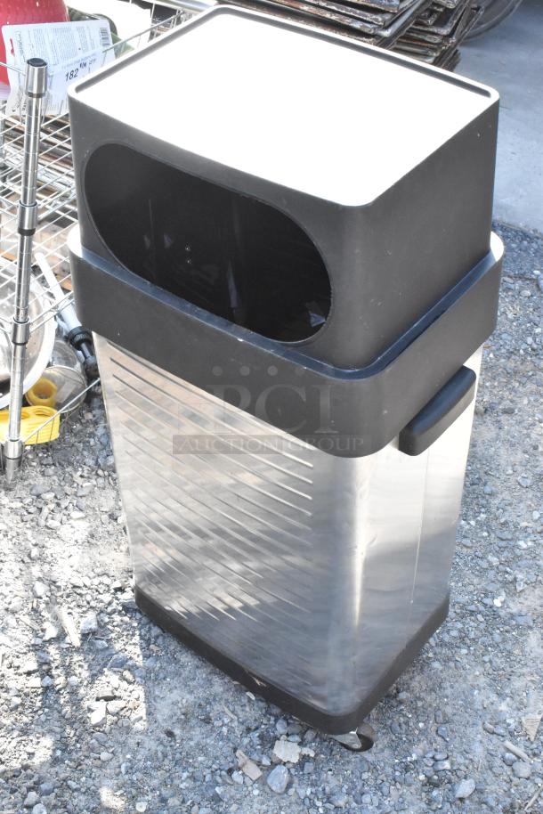 Stainless steel trash can with black plastic lid and wheels, shown outdoors, displaying its modern design and durable construction.