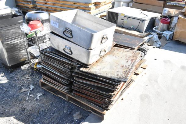 Lot of metal roasting pans and perforated baking sheets stacked on a pallet, showing signs of wear, ideal for bulk purchase.