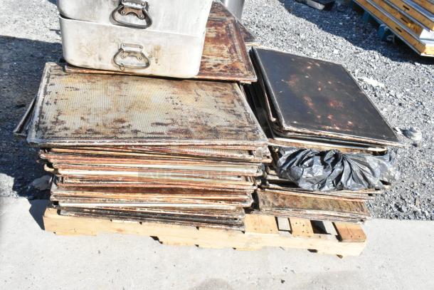 Lot of metal roasting pans and perforated baking sheets, used condition, stacked on a wooden pallet outdoors.