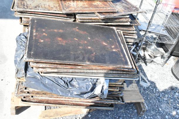 Lot of metal roasting pans and perforated baking sheets in used condition, stacked on a wooden pallet outdoors.