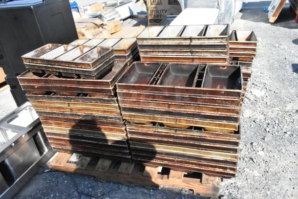 Lot of stacked metal 4-loaf baking pans, showing wear and rust, on a wooden pallet outdoors.