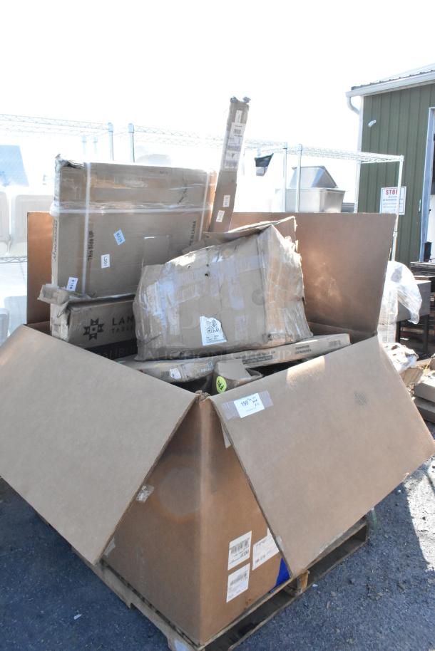 Large boxes of new scratch and dent items, including a stainless steel table and Regency posts, stacked on a pallet.
