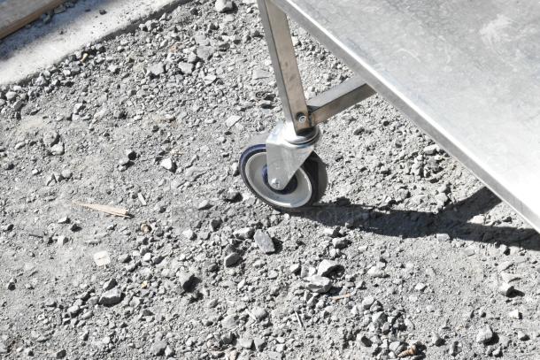 Stainless steel cart with under shelf on commercial casters, showing robust wheel and durable construction on gravel surface.